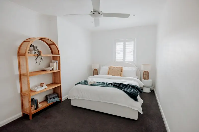 Soft white bedroom with rattan shelving, plantation shutters, and layered linens.
